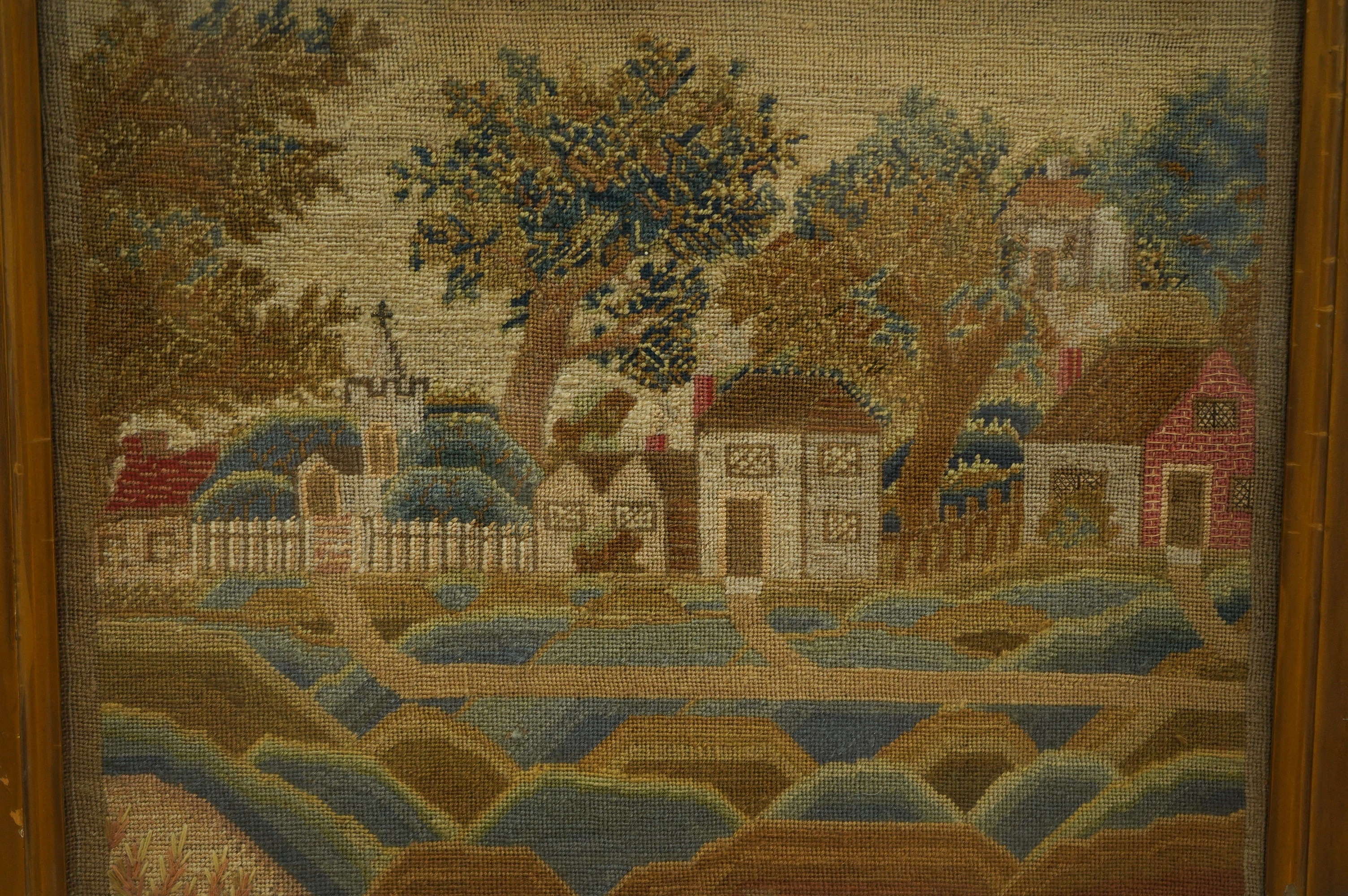 A framed 19th century woolwork of a village scene of cottages, houses, a church amongst trees and a picket fence with paths leading to another, within an undulating foreground, the leaves of the trees worked in various s